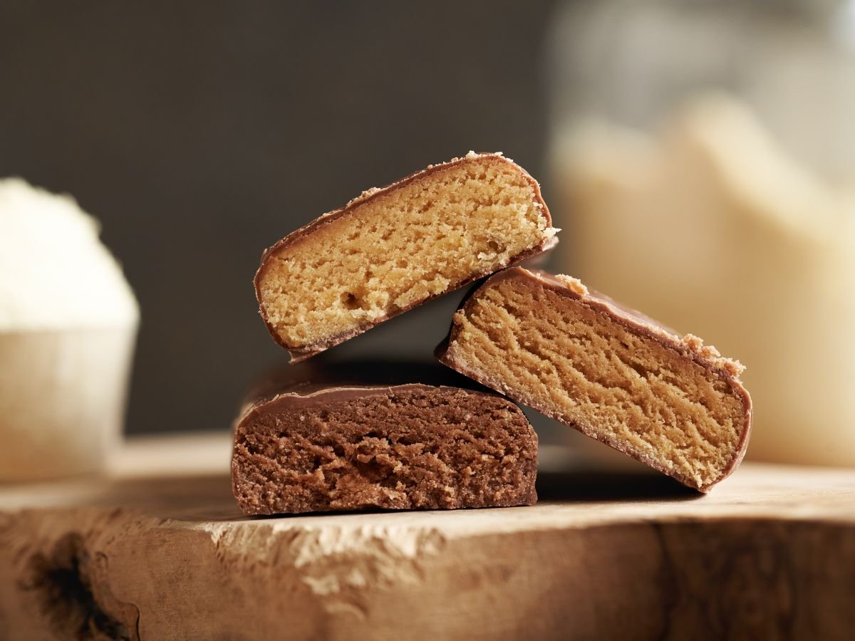 Three chocolate covered protein bar flavors cut in half on a cutting board with protein powder in the background.