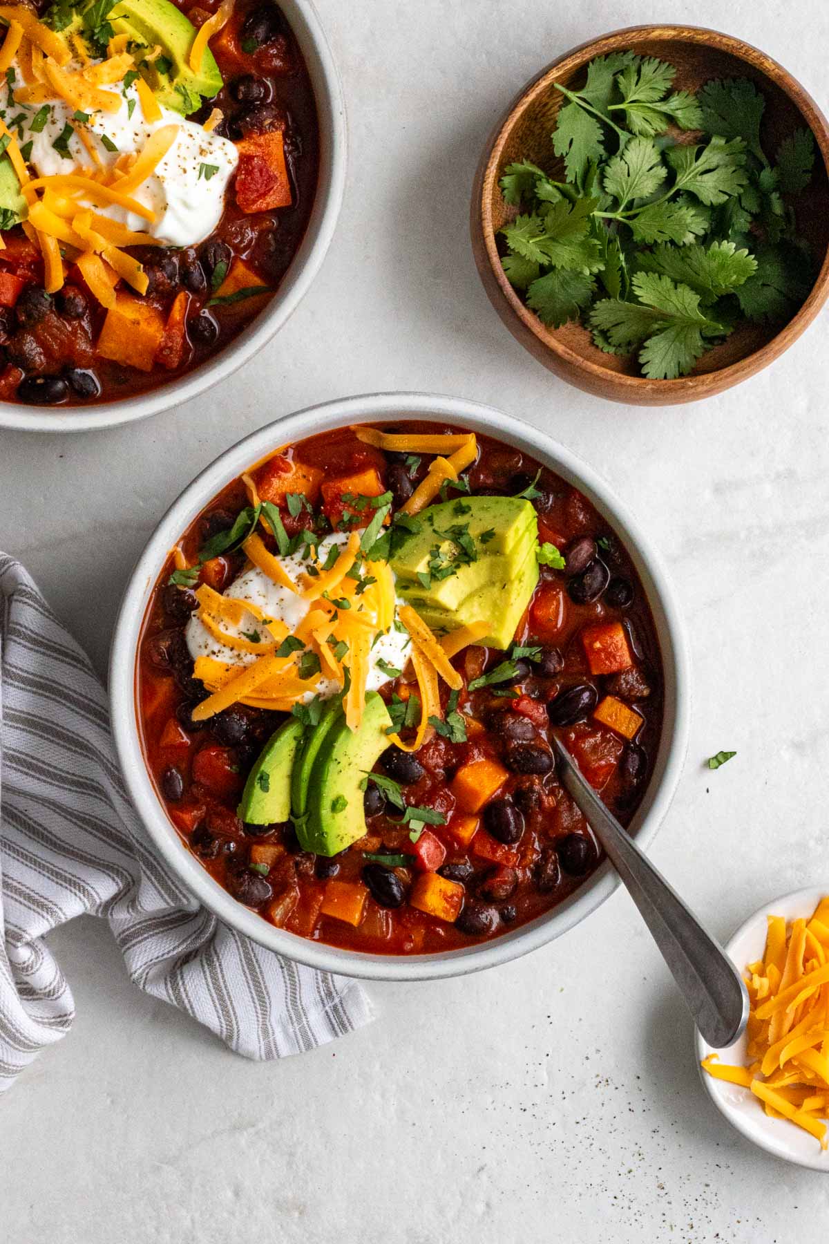 Two bowls of black bean and sweet potato chili with sour cream, sliced avocado, grated cheese, and cilantro on top with a spoon and tea towel on the side on a white background.