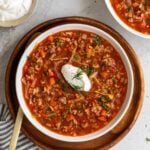 Two bowls of cabbage roll soup with sour cream and fresh dill on top on a wood platter with a gold spoon and a striped tea towel.