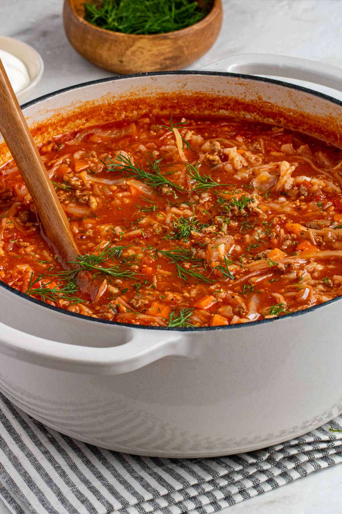 Large white pot with cabbage roll soup with a wood serving spoon and chopped dill on top.