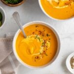 Two bowls of carrot ginger coconut soup with a drizzle of coconut milk and chopped cilantro on top with a side of bread and cilantro on a white background.