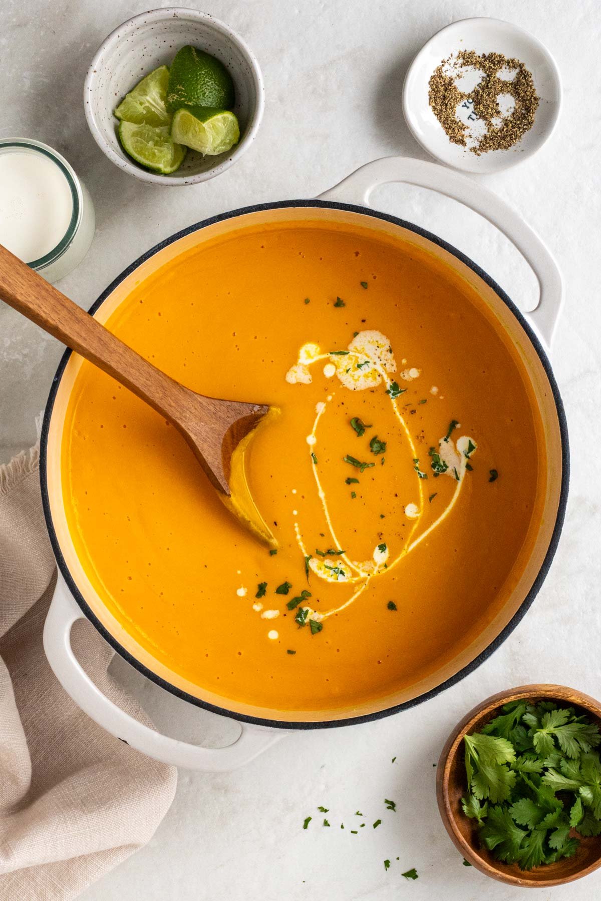 Large pot of ginger coconut carrot soup with a wooden spoon and a tea towel on the side with cilantro, lime wedges, coconut milk, and black pepper on the side on a white background.