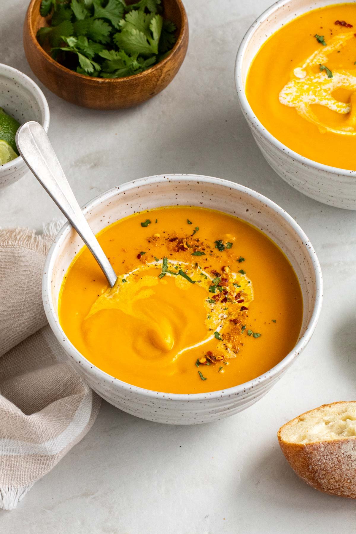 Two bowls of ginger carrot soup with a side of cilantro, lime wedges, and crusty bread.