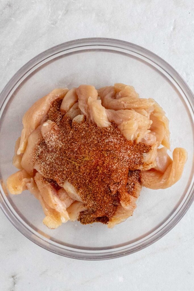 Bowl with chicken strips with chili powder, paprika, cumin, garlic powder, cayenne pepper, and salt on top.