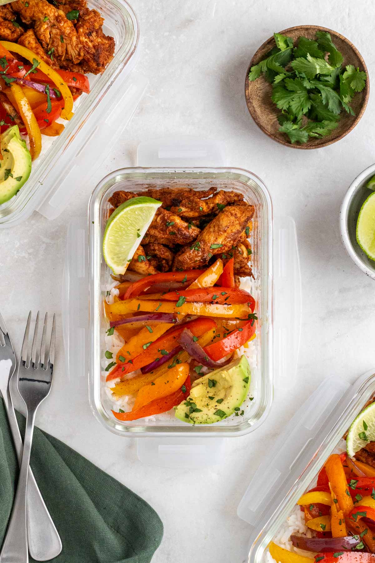 Three chicken fajita meal prep bowls with a fork and tea towel and a side of cilantro and lime wedges.