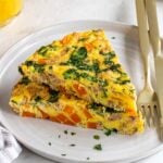Plate with two slices of sweet potato and sausages frittata with a glass of orange juice, parsley, and crusty bread in the background.