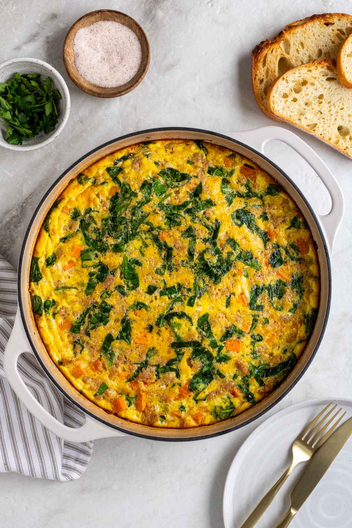 SKillet with sweet potato and sausage frittata with salt, parsley, and bread on the side with a plate with gold fork and knife and a tea towel.