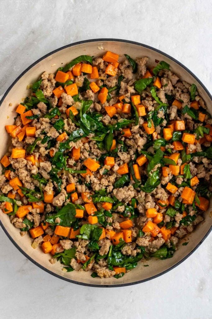 Skillet with sautéed onion, browned sausage, diced sweet potatoes, and wilted spinach.