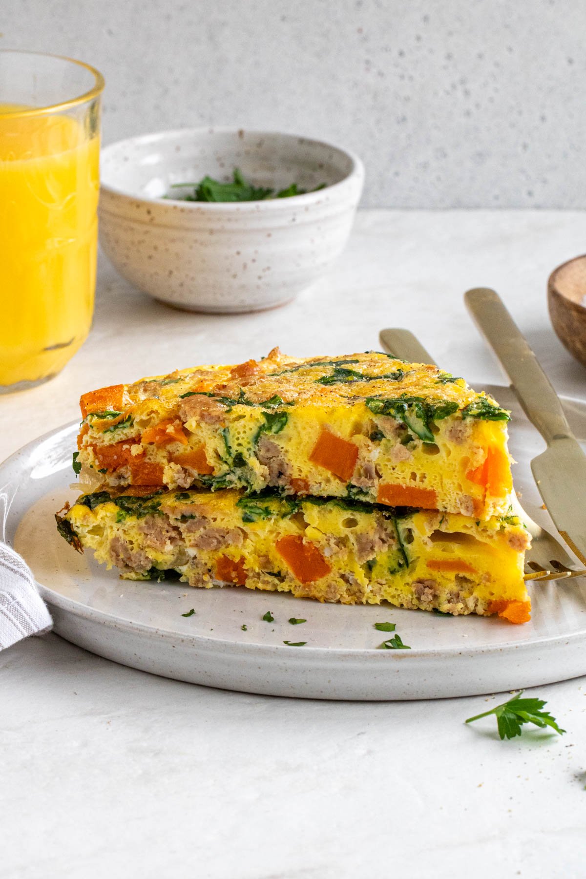 Close up of two slices of sweet potato and sausage frittata on a white plate with gold utensils with a small glass of orange juice and parsley in the background.