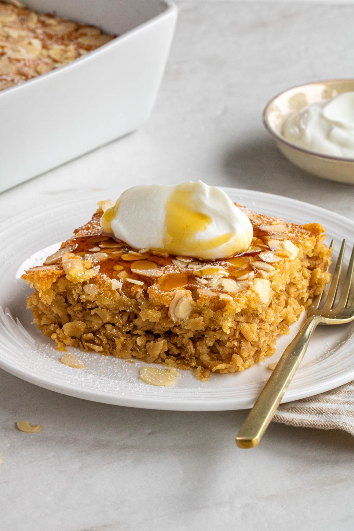 Slice of almond croissant baked oats on a white plate with Greek yogurt and maple syrup on top with a gold fork.