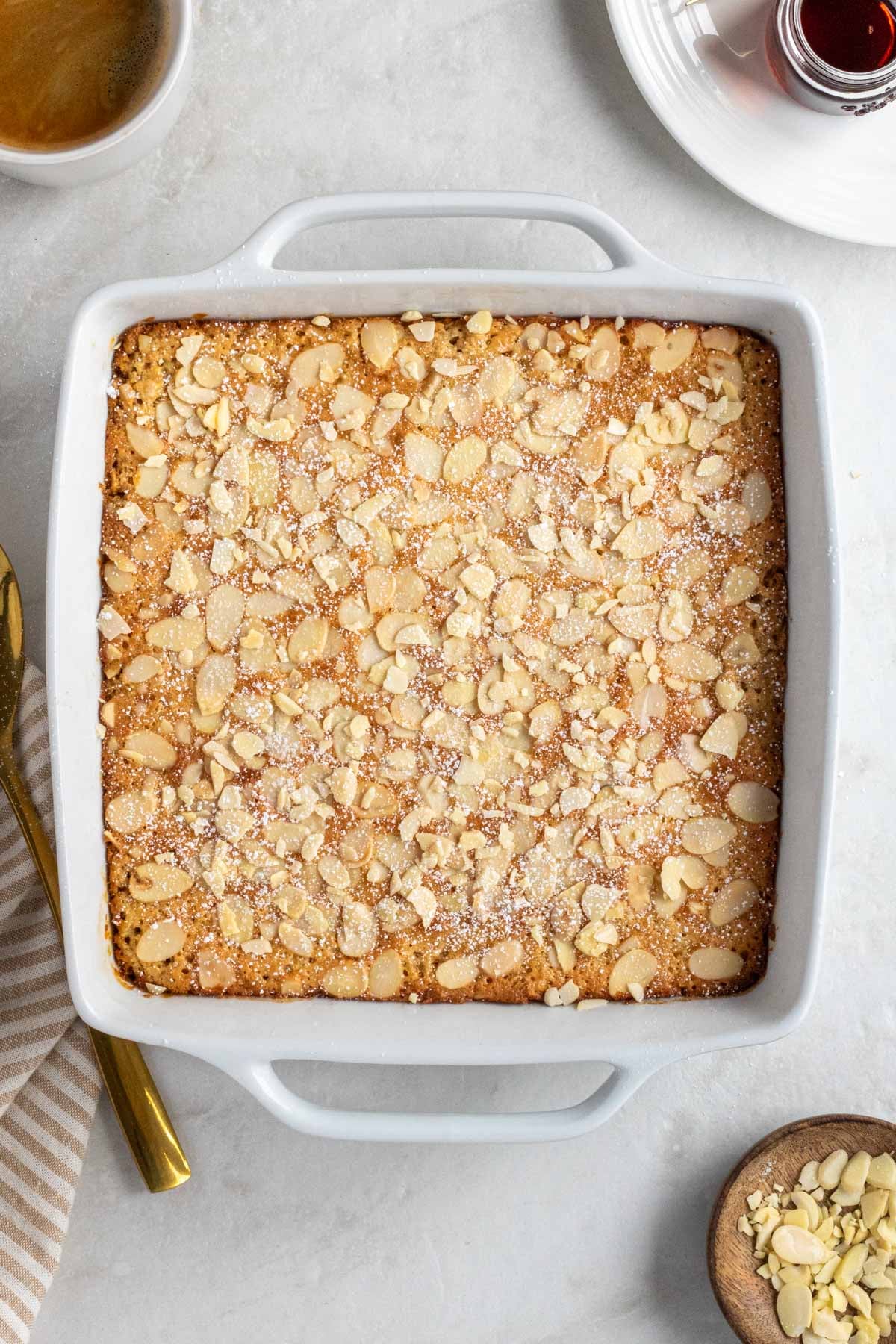 White baking dish of almond croissant baked oats with powdered sugar on top with a beige striped tea towel, gold serving spoon, wood bowl of flaked almond, maple syrup, and coffee on the side.