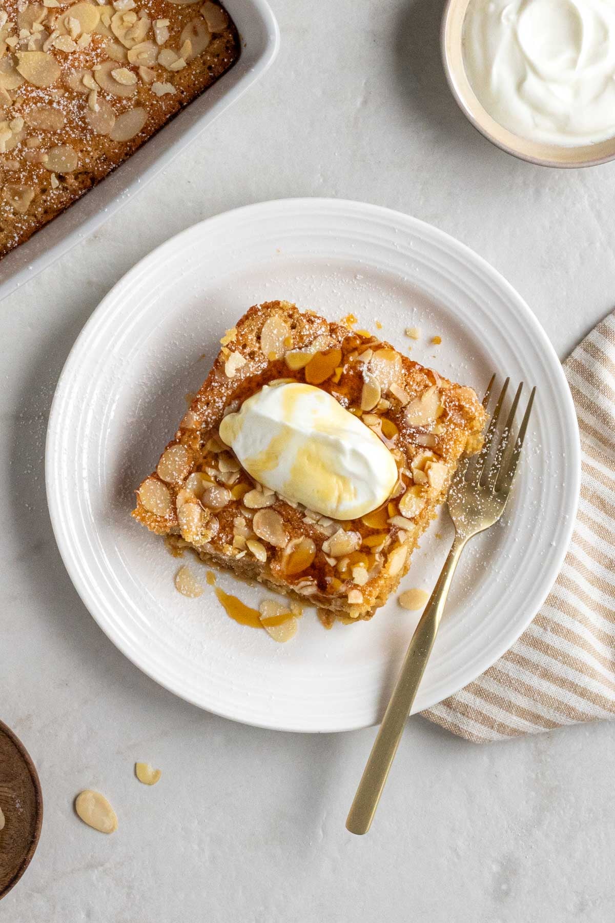 White plate with a slice of baked almond croissant oatmeal with powdered sugar, greek yogurt, and maple syrup on top and flaked almond on the side. 