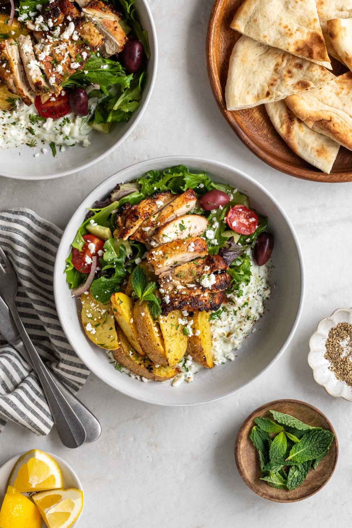 Two chicken tzatziki bowls on a white background with lemon wedges, fresh mint, black pepper, and pita bread on a white background.