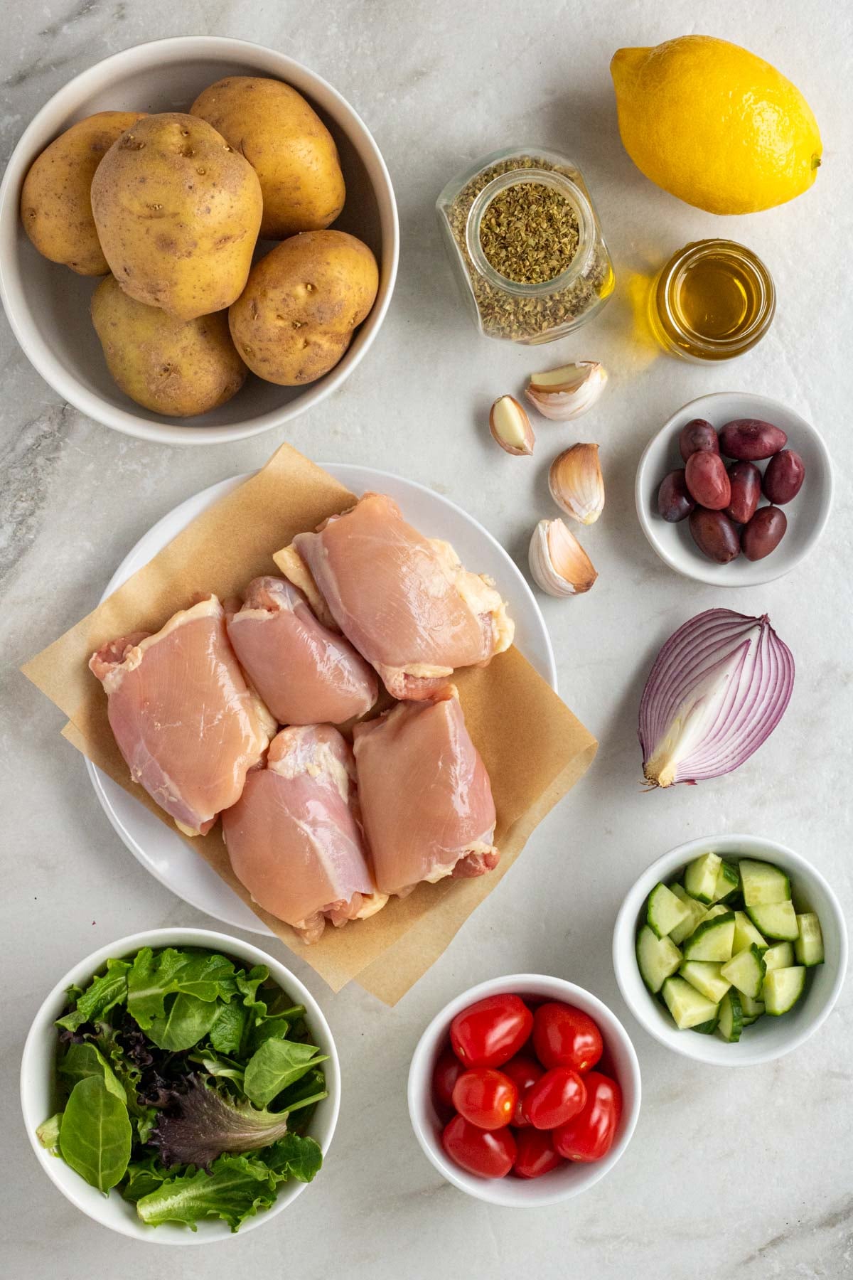 Ingredients chicken tzatziki bowls: yellow potatoes, chicken thighs, lemon, olive oil, oregano, garlic, kalamata olive, red onion, cucumber, grape tomatoes, leafy greens.