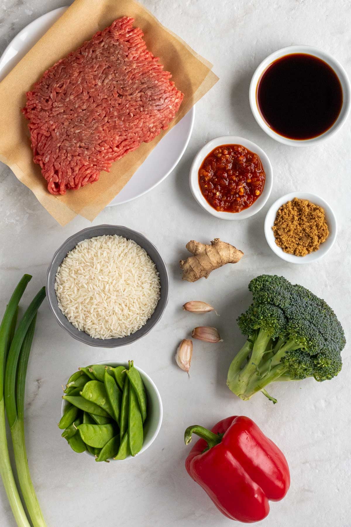 Ingredients for ground beef stir fry: white rice, extra lean ground beef, green onions, red bell pepper, broccoli florets, garlic, ginger, soy sauce, chili paste, sesame oil, rice vinegar, brown sugar, and sesame seeds.