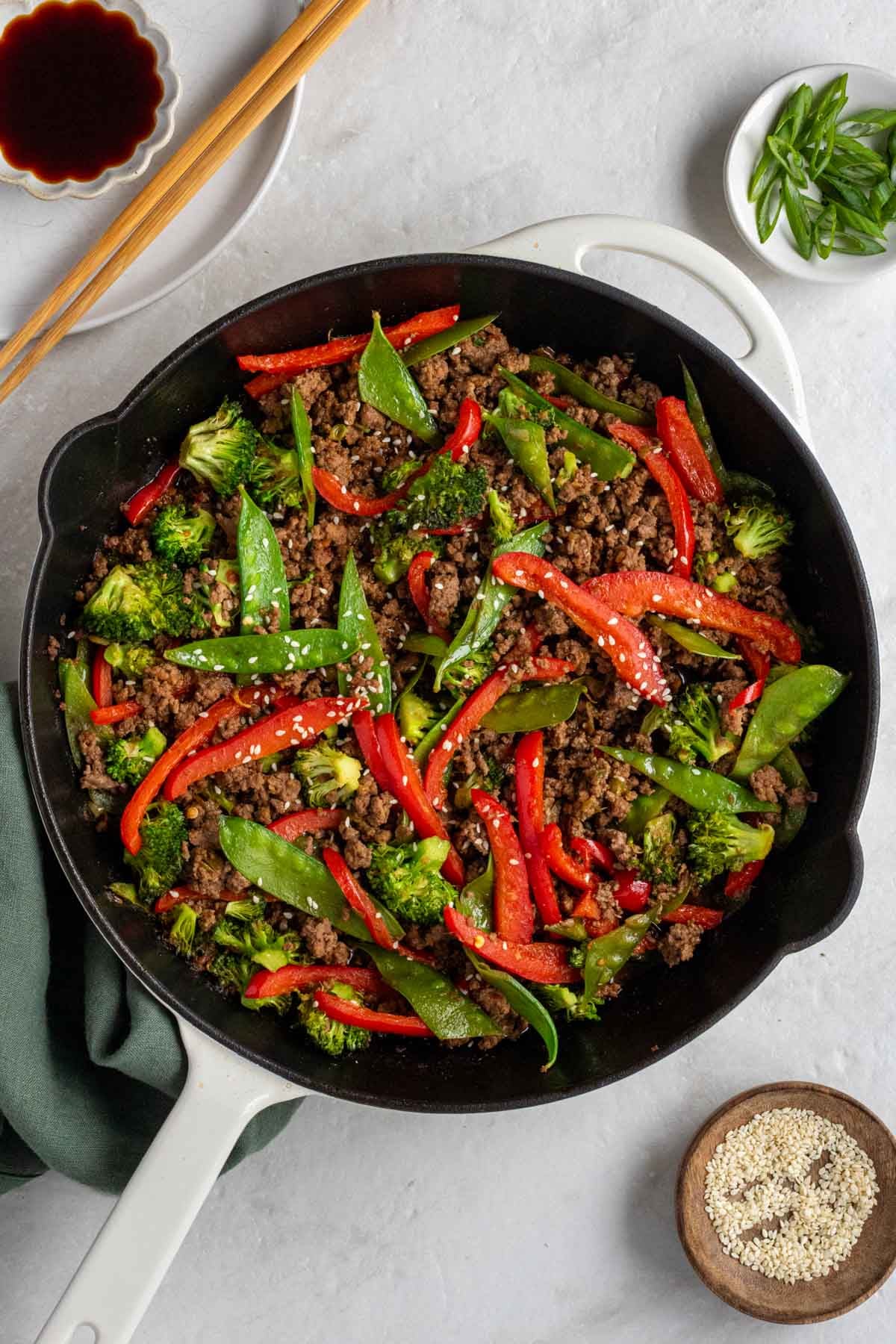 Pan with ground beef stir fry with a green tea towel with sesame seeds, soy sauce, and green onions on the side on a white background.