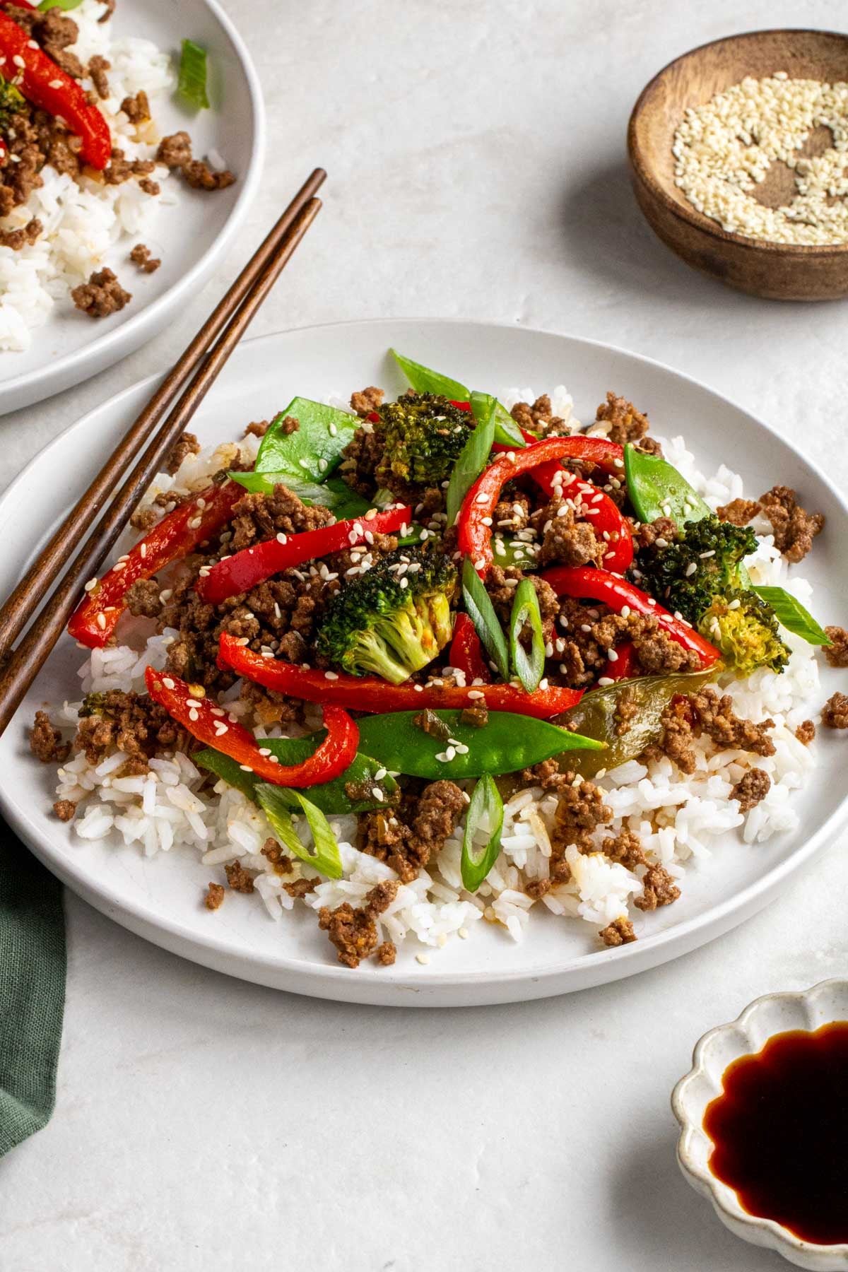 Two plates of easy ground beef stir fry with homemade stir fry sauce on a white background with wood chopsticks and sesame seeds in a wooden bowl.