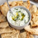 Close up of a small bowl of homemade tzatziki sauce with olive oil and dill on top with a serving spoon on a plate with pita bread.