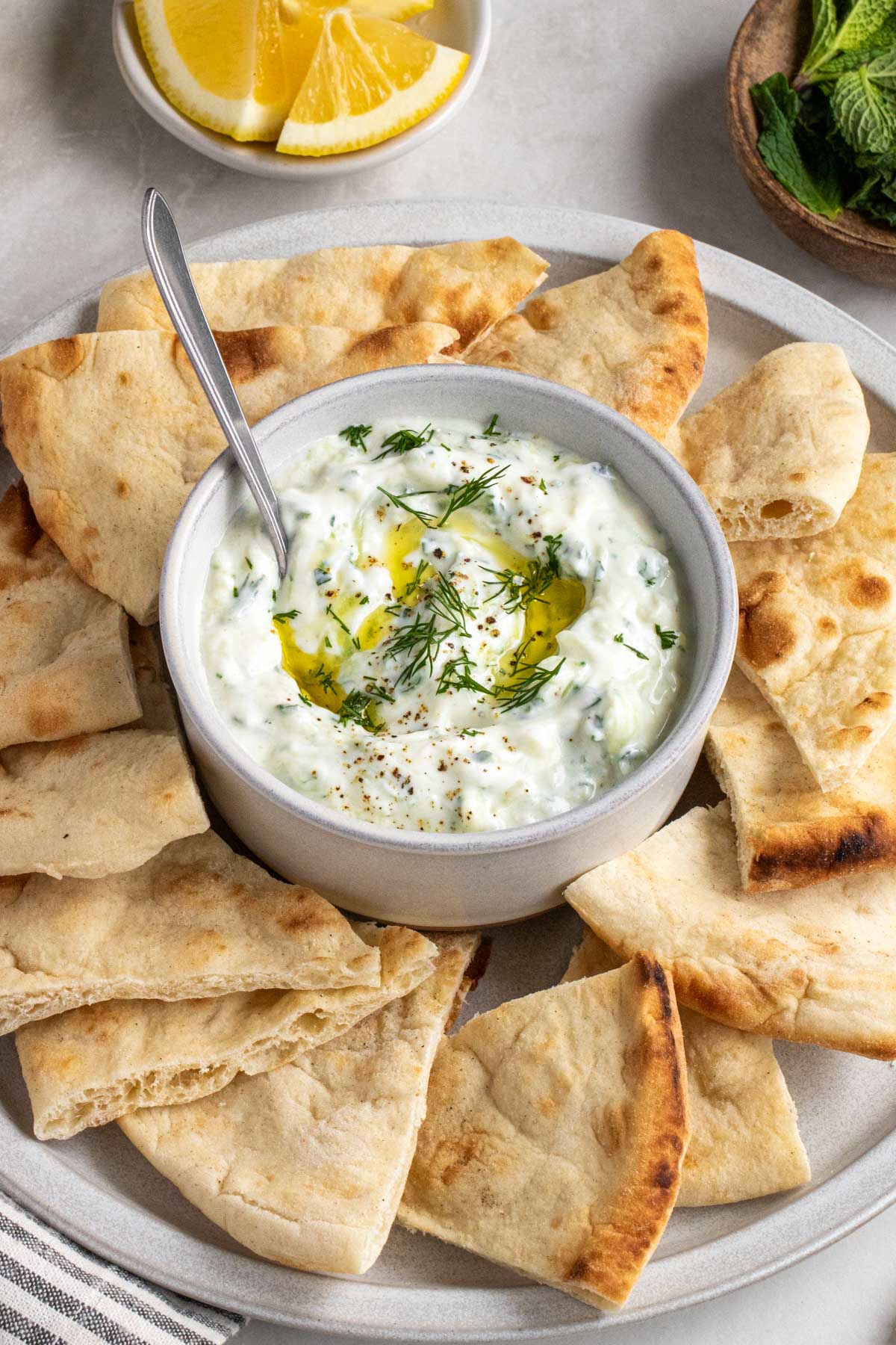 Close up of a small bowl of homemade tzatziki sauce with olive oil and dill on top with a serving spoon on a plate with pita bread.