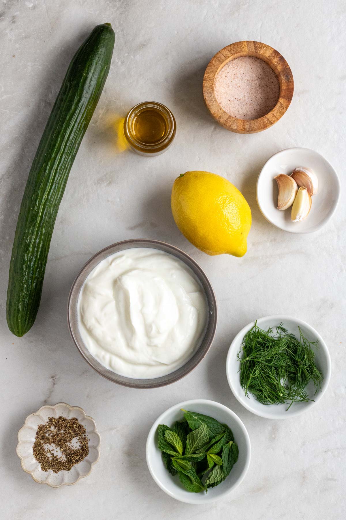 Ingredients for homemade tzatziki sauce: Greek yogurt, English cucumber, garlic, lemon, olive oil, fresh mint, fresh dill, salt, and black pepper.