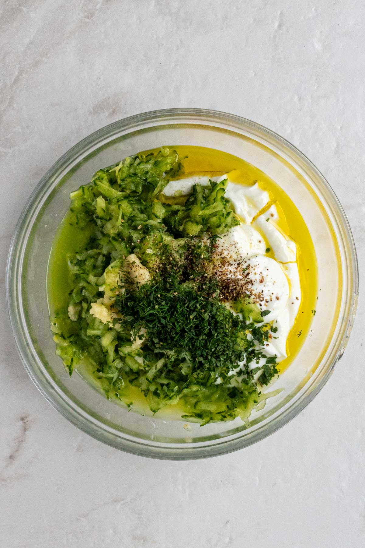 Glass bowl with Greek yogurt, shredded cucumber, grated cucumber, olive oil, lemon juice, chopped dill, chopped mint, salt, and black pepper.