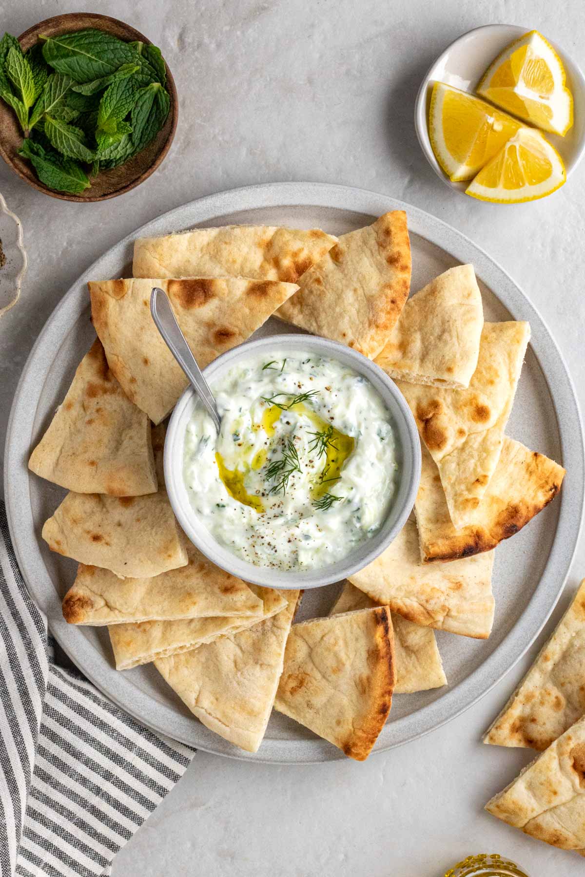 Platter with pita chips with tzatziki with lemon wedges and fresh mint on the side on a white background.