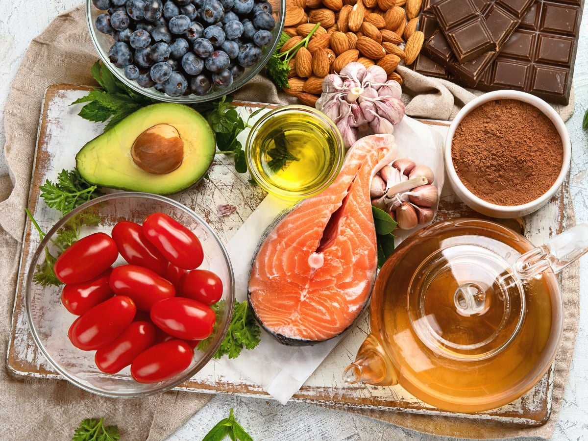 Blueberries, almonds, dark chocolate, garlic, cocoa powder, olive oil, avocado, tomatoes, and salmon on a white cutting board with a beige tea towel.