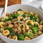 Close up of a white bowl with sausage and rapini pasta freshly grated parmesan cheese on top with a gold fork and a wood cutting board and glass of water in the background.