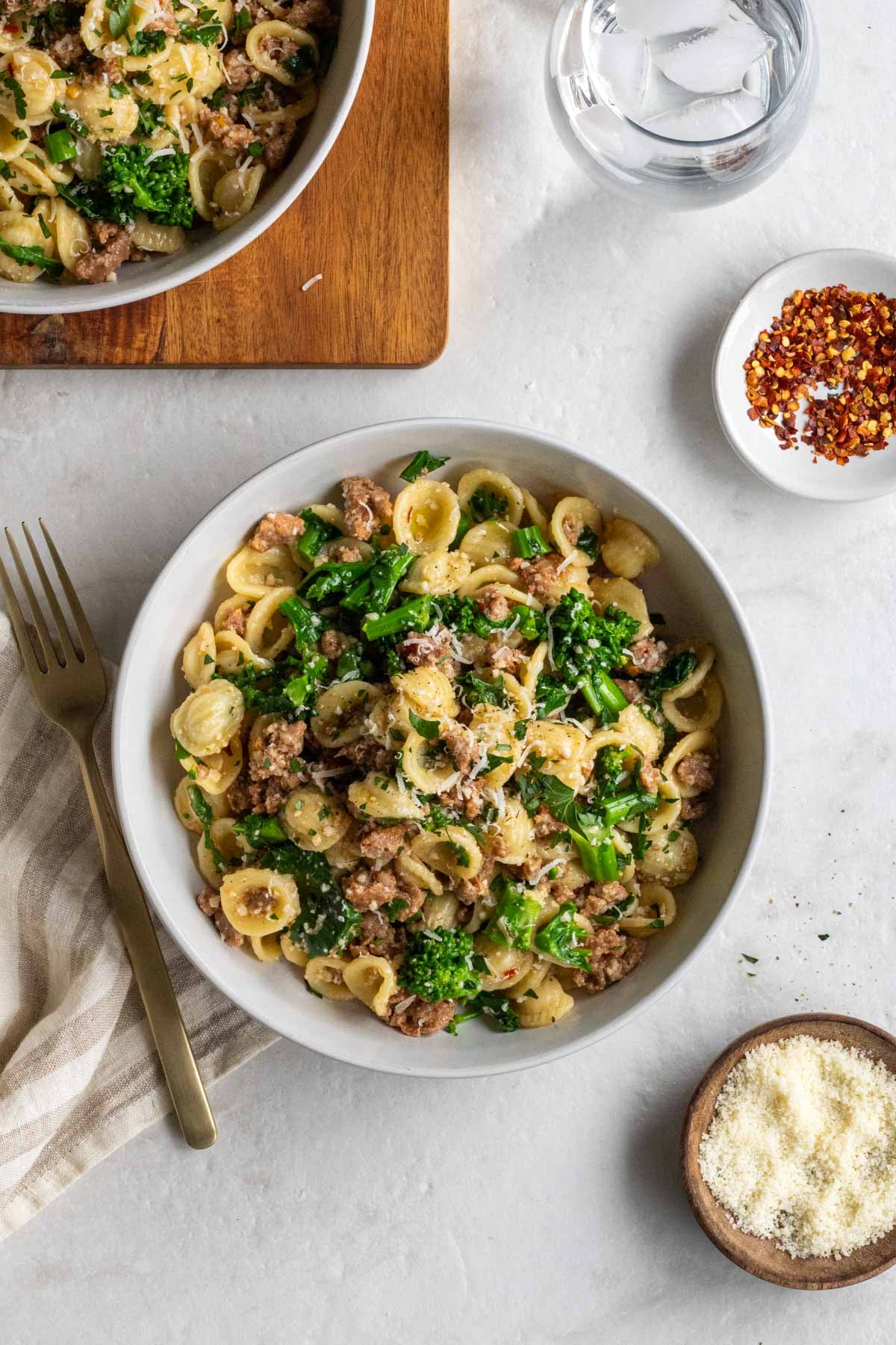 Two bowls of sausage and broccoli rabe pasta with a fork, tea towel, glass of water, and red pepper flakes on the side.