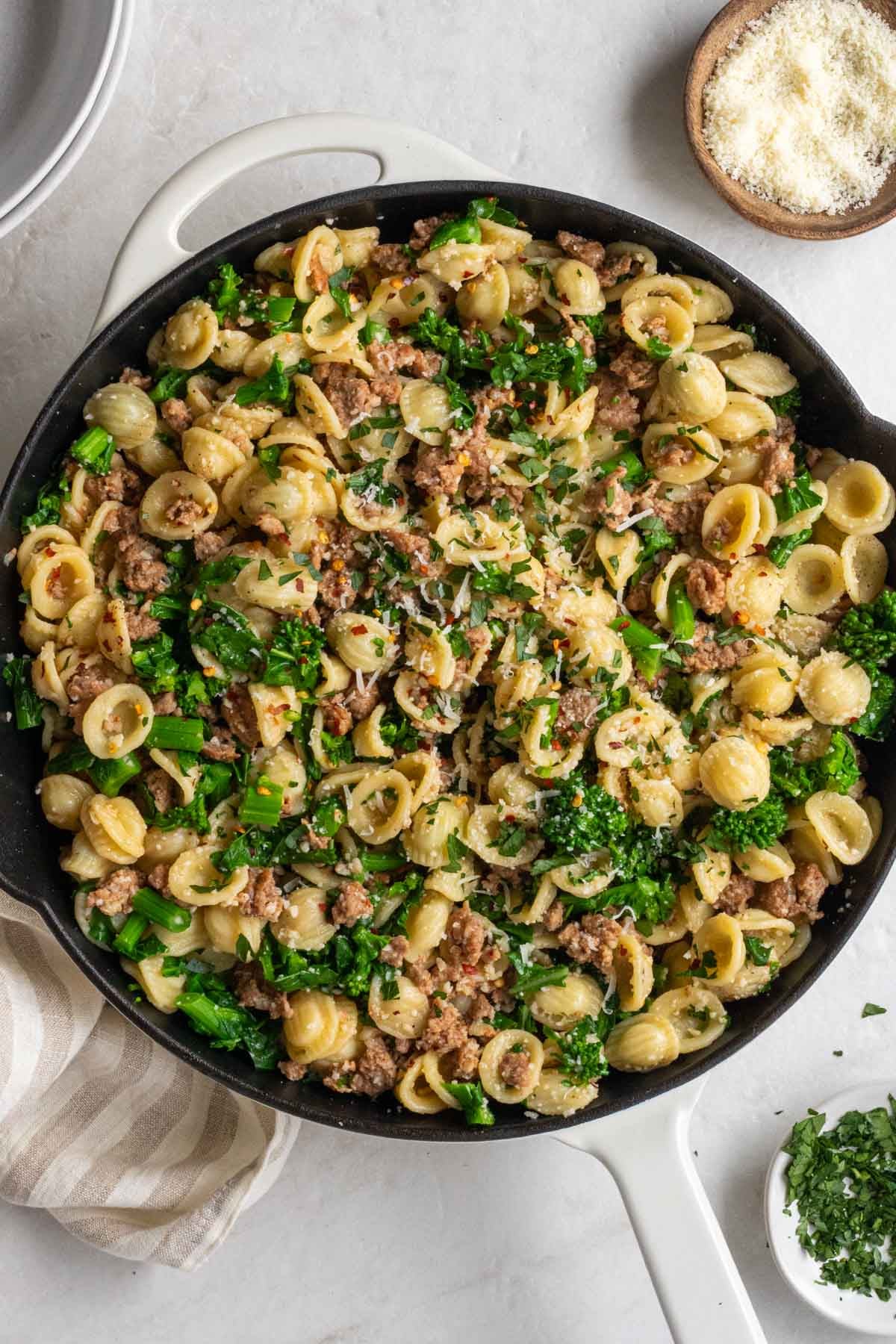 Large cast iron pan with rapini sausage pasta with a small wood bowl of pecorino cheese on the side.
