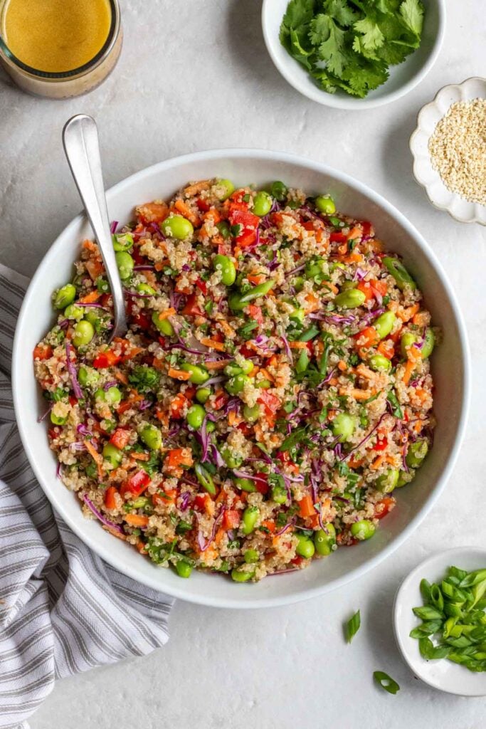 Large white bowl with quinoa edamame salad with a silver serving spoon with a jar of ginger sesame dressing on the sie with a bowl of chopped cilantro and sesame seeds on a white background.
