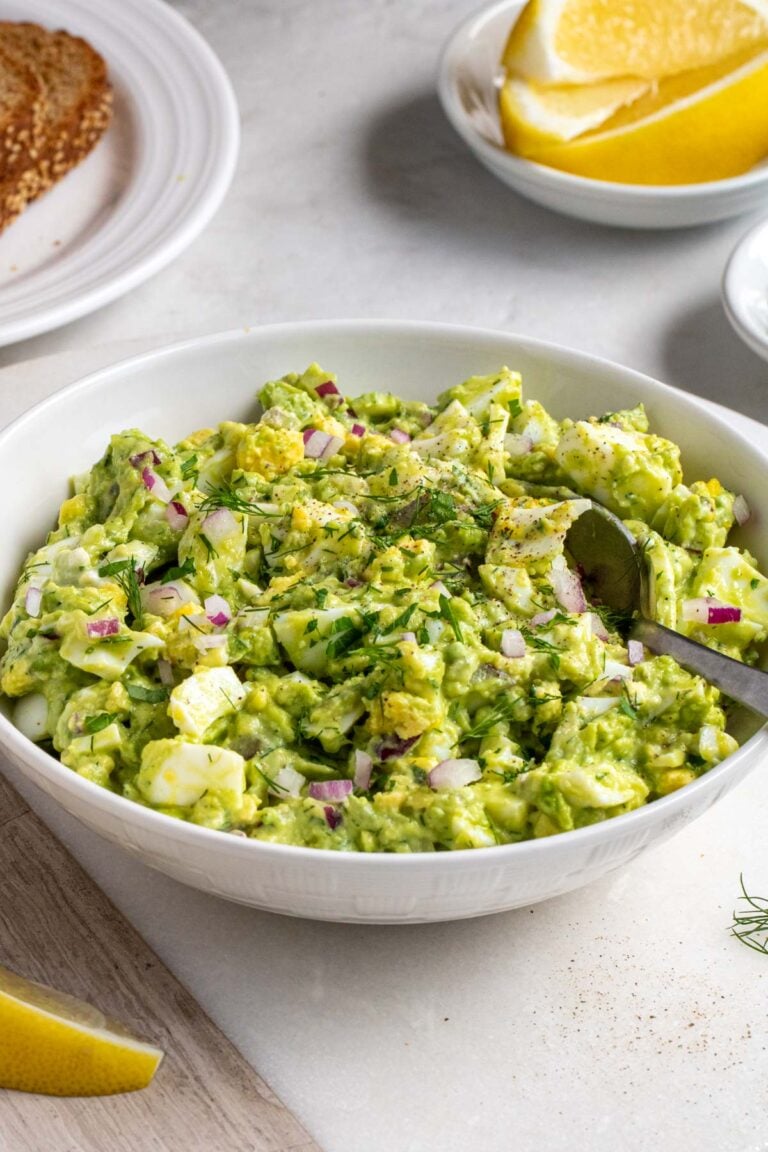 White bowl of avocado egg salad with a silver spoon on a white background with lemon wedges and whole grain bread in the background.