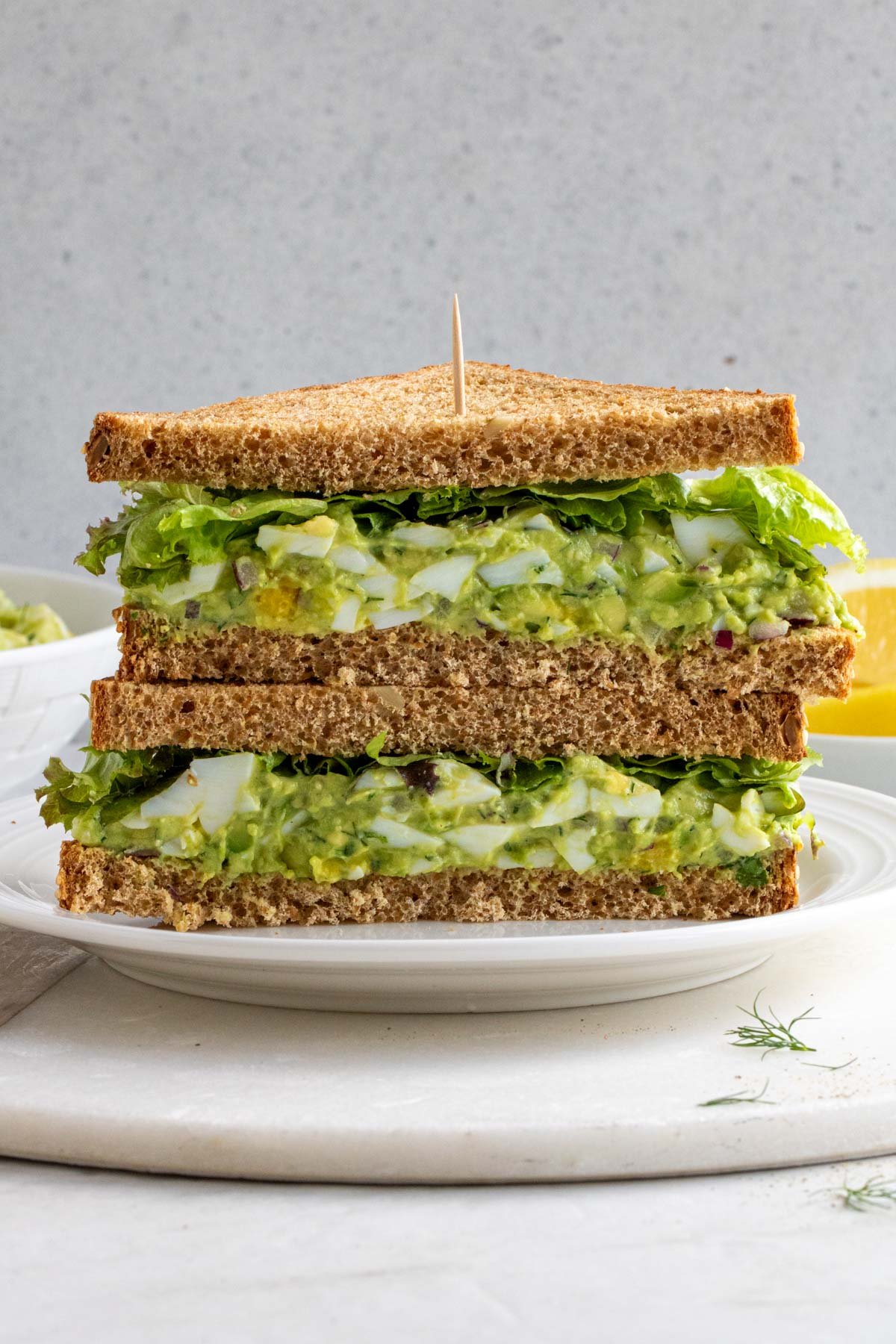 Stacked and sliced avocado egg salad sandwich with lettuce with a toothpick on a white plate with fresh dill and lemon wedges on the side on a white background.