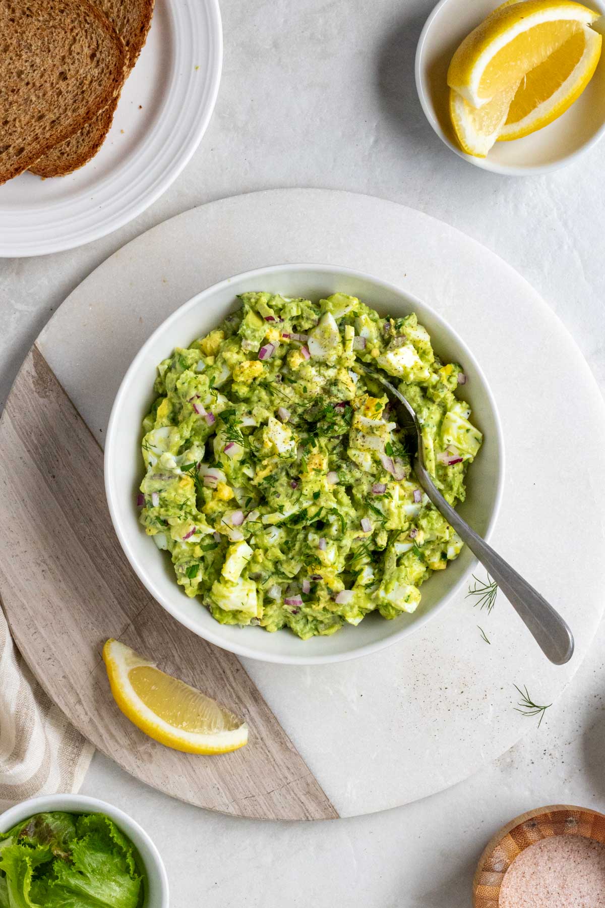 Overhead of a white bowl of avocado egg salad on a white cutting board with a serving spoon with whole grain bread, lemon slices, salt, and lettuce on the side.
