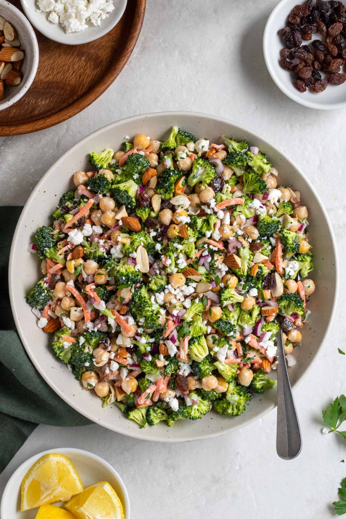 Large bowl of broccoli and chickpea salad with a silver serving spoon and a green tea towel on a white background with small bowls of almonds, feta cheese, and raisins on the side.