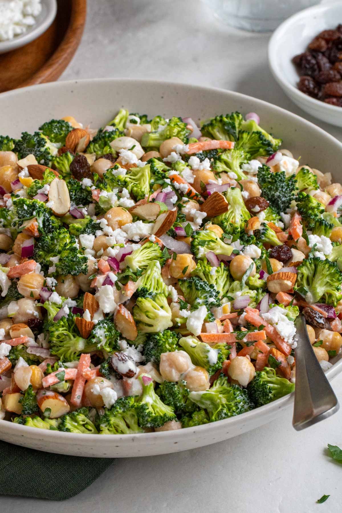 Close up of a beige bowl broccoli chickpea salad with a serving spoon with a small bowl of raisins in the background.