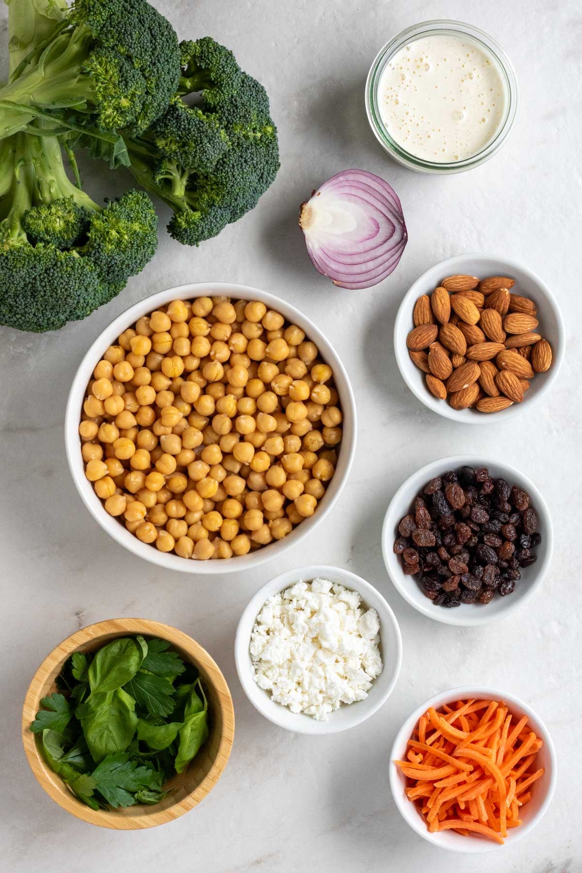 Head of broccoli, red onion, and bowls of creamy dressing, almonds, chickpeas, raisins, feta cheese, fresh herbs, and shredded carrots on a white background.