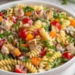 Close up of a large white bowl with creamy chicken pasta with arugula and salad dressing in the background and a wooden serving spoon on the side.