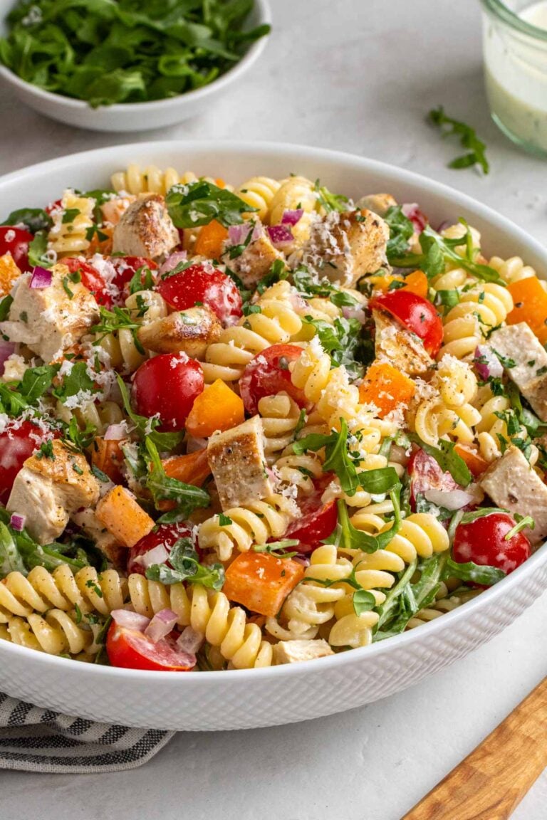 Close up of a large white bowl with creamy chicken pasta with arugula and salad dressing in the background and a wooden serving spoon on the side.