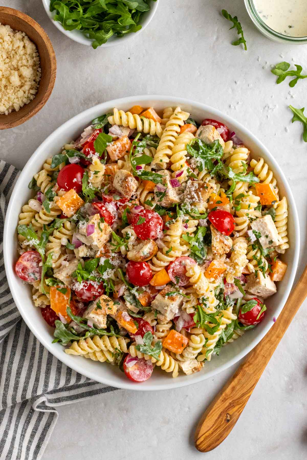 White bowl of creamy chicken pasta salad with a jar creamy salad dressing, a small bowl of arugula, and wooden bowl of parmesan, a wooden serving spoon, and tea towel on the side.