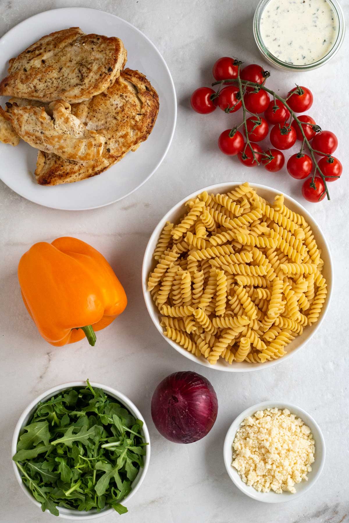 Plate of cooked chicken breasts with bowls of fusilli pasta, grape tomatoes, arugula, and parmesan, with a red onion and orange bell pepper, and a bowl of creamy salad dressing on a white background.
