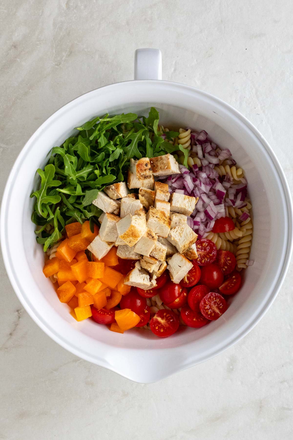 Large bowl with cooked fusilli pasta, chopped cooked chicken, sliced cherry tomatoes, diced orange bell pepper, sliced red onion, chopped arugula, and freshly grated parmesan cheese.