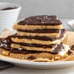 Stack of peanut butter frozen yogurt bark on a white plate with a bowl of melted chocolate and bowl of chopped peanuts in the background.