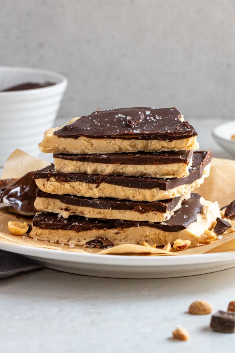 Stack of peanut butter frozen yogurt bark on a white plate with a bowl of melted chocolate and bowl of chopped peanuts in the background.
