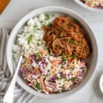 Close up of a slow-cooker BBQ pulled pork bowls white rice and coleslaw with chopped cilantro on top with a fork and white and grey tea towel on white background.