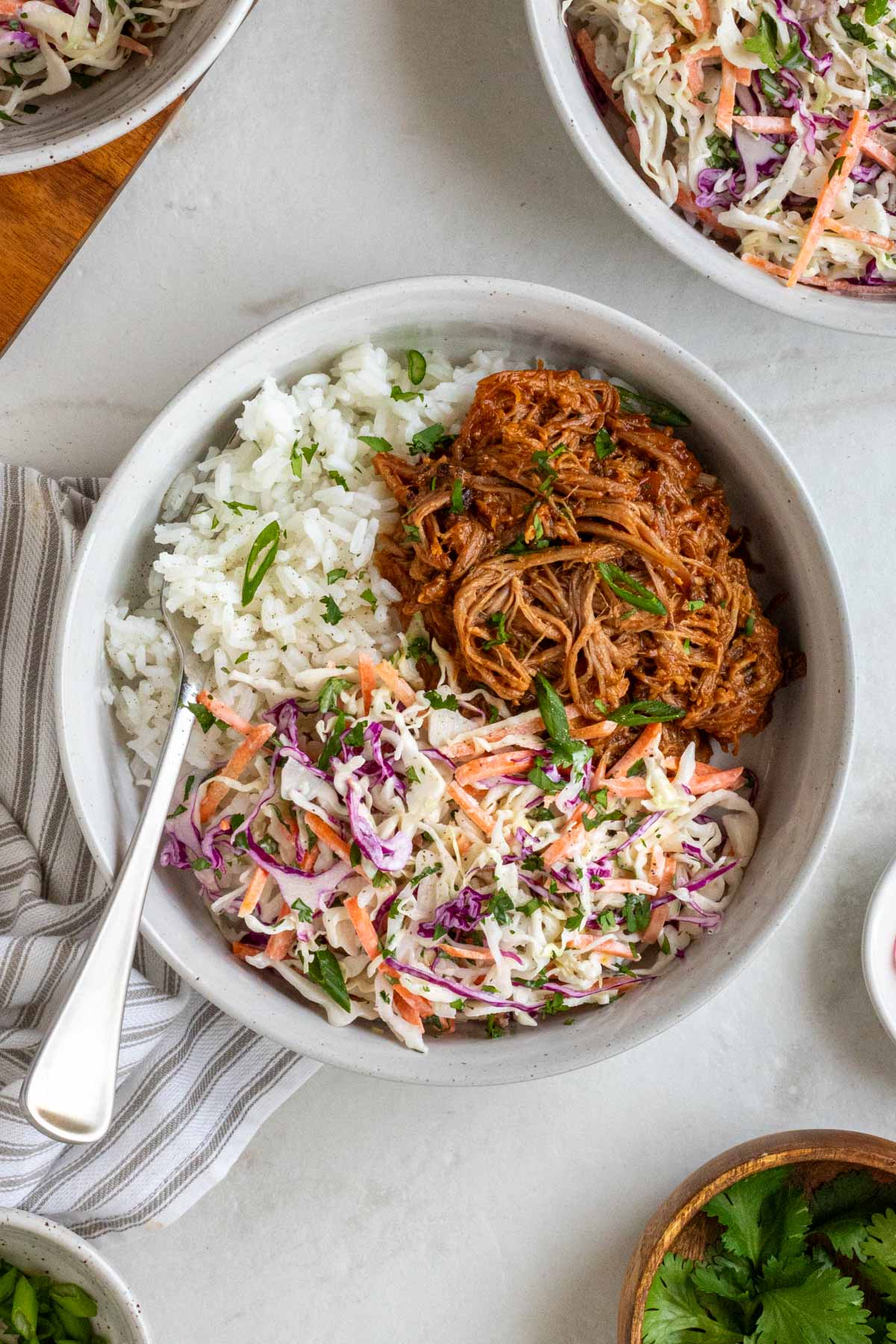 Close up of a slow-cooker BBQ pulled pork bowls white rice and coleslaw with chopped cilantro on top with a fork and white and grey tea towel on white background.