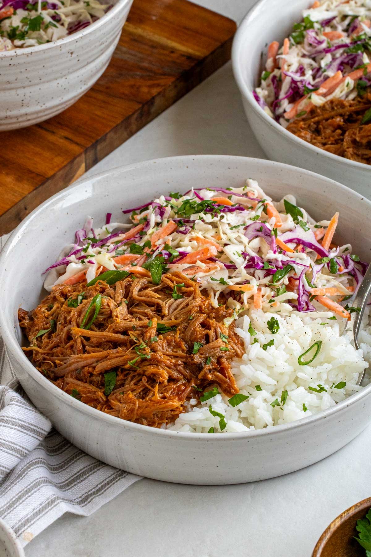 Two slow-cooker pulled pork bowls on a white background with a bowl of creamy coleslaw on a wood cutting board on the side.