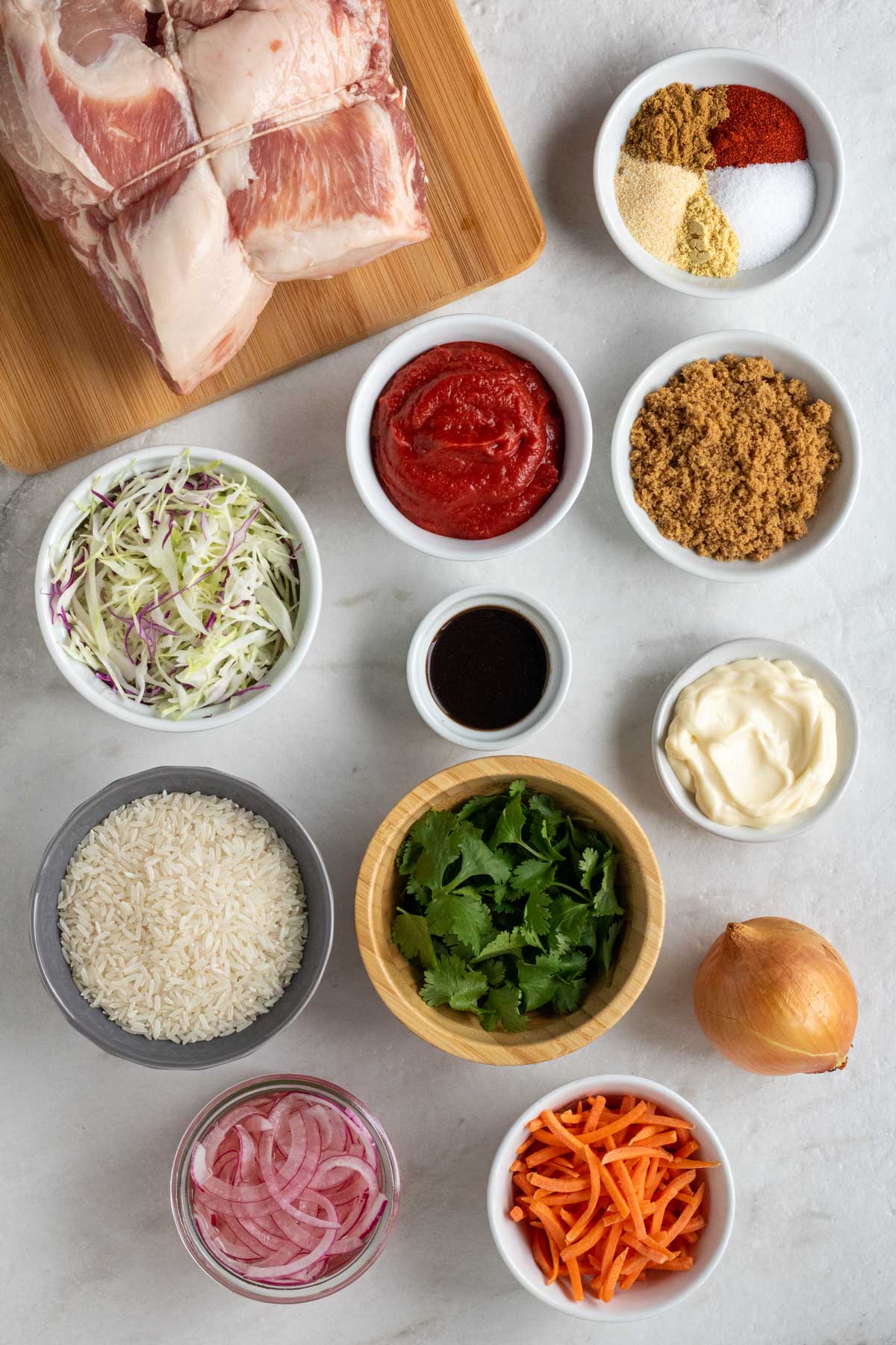 Pork shoulder on a wood cutting board with bowls of spices, tomato paste, brown sugar, worcestershire sauce, mayonnaise, shredded cabbage, white rice, cilantro, white onion, shredded carrots, and pickled onions on a white background. 