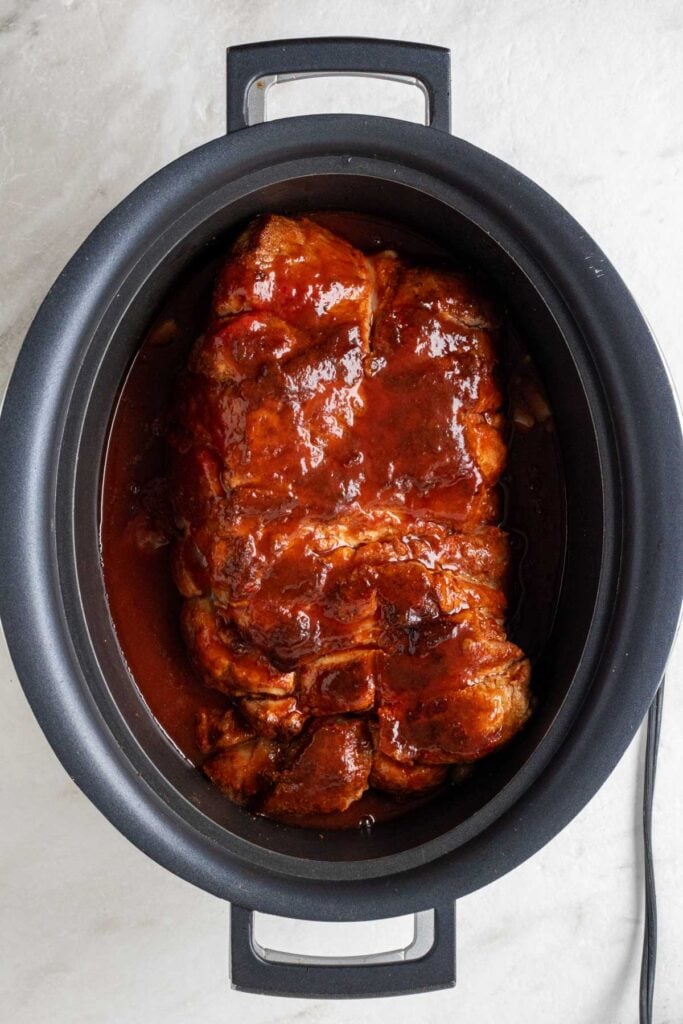 Seared pork shoulder in a slow cooker covered in barbecue sauce.