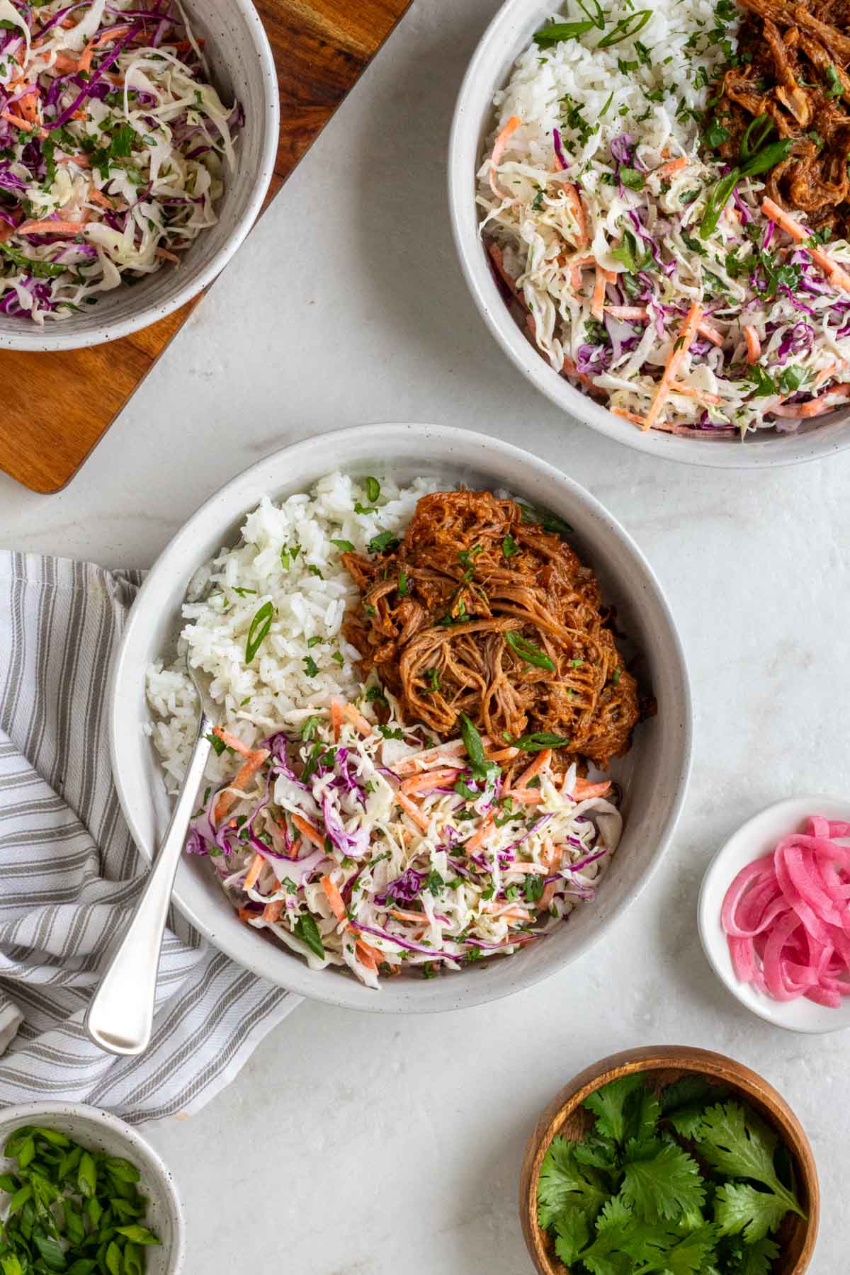 Two BBQ pulled pork bowls with white rice and creamy coleslaw on a white background with a bowl of coleslaw, pickled onion, cilantro, and green onions on the side.